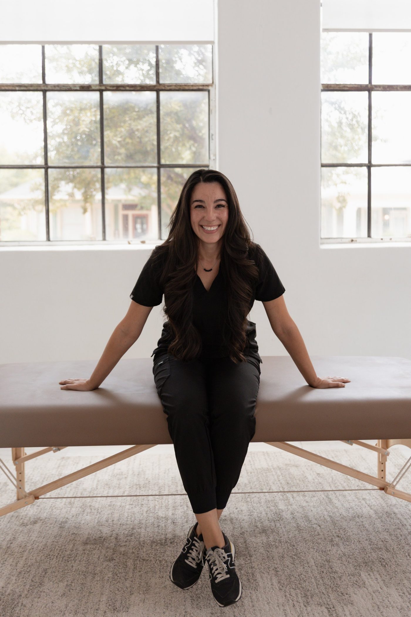 Valerie sitting on the edge of a massage table in front of tall windows